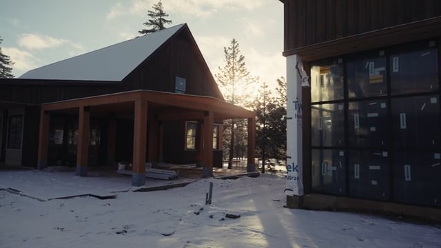 A modern house under construction in a snowy landscape, rooted in Suncadia Social living. Sunlight filters through trees, highlighting dark wood siding and large windows, while building materials are scattered on the ground.
