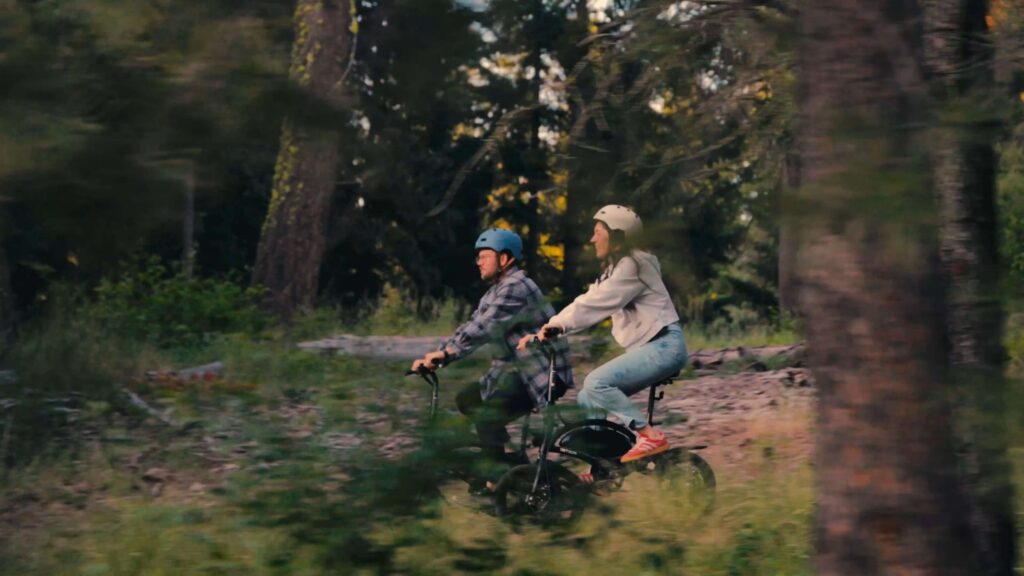 Two people wearing helmets ride bicycles side by side on a dirt path through a forest, surrounded by tall trees and greenery, enjoying a day Rooted In Suncadia Social adventure.