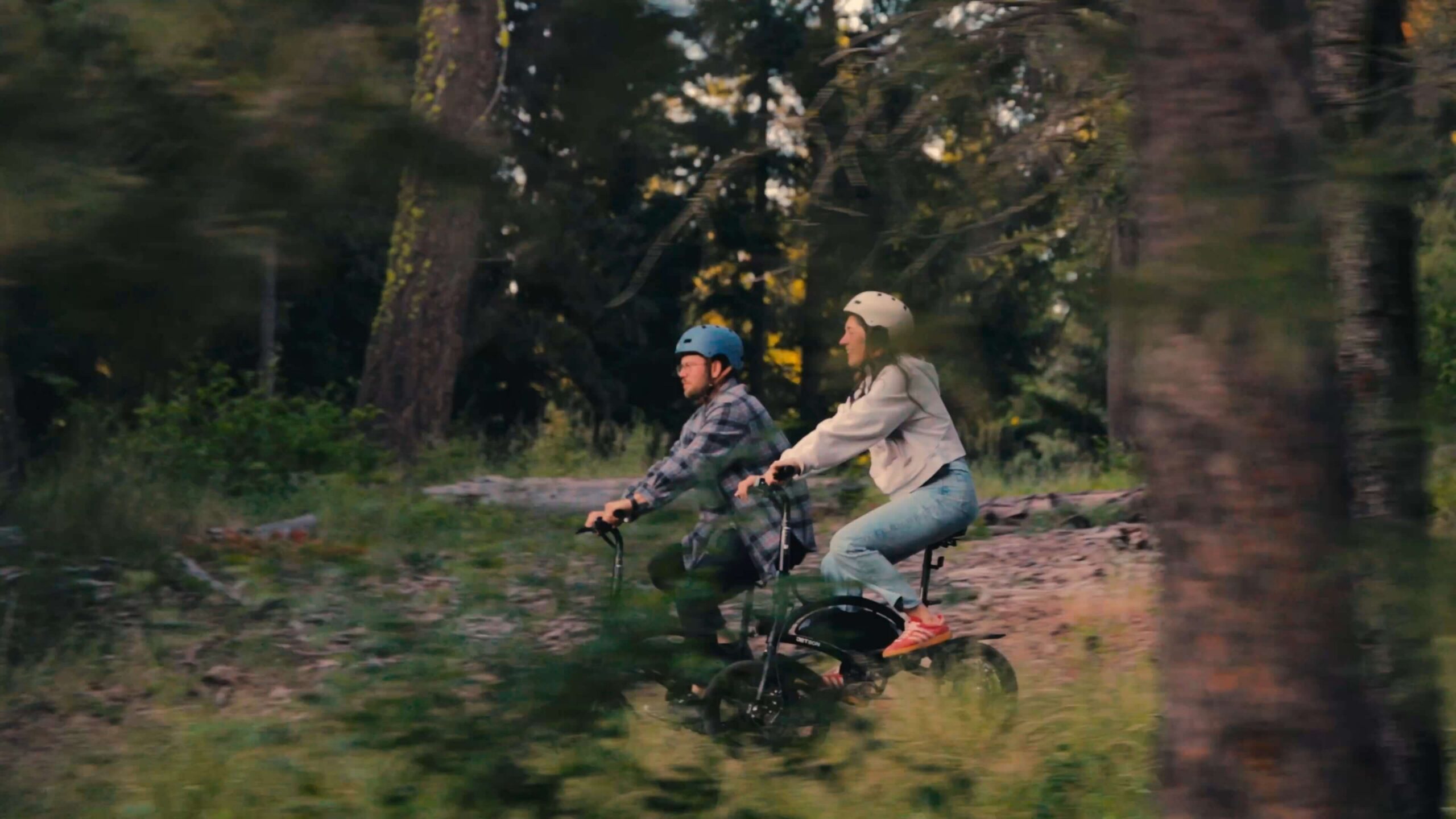 Two people wearing helmets ride bicycles side by side on a dirt path through a forest, surrounded by tall trees and greenery, enjoying a day Rooted In Suncadia Social adventure.