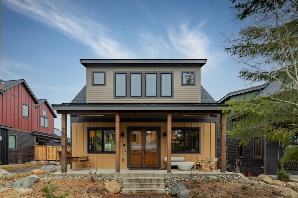 A modern two-story house with beige and gray siding, a covered front porch with wooden posts, large windows, outdoor seating, and a landscaped front yard with rocks and small trees under a blue sky.