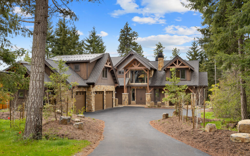 A large, modern rustic house with wooden and stone accents sits among tall trees. The driveway leads to a three-car garage and the entrance features exposed timber beams and a balcony above the front door.