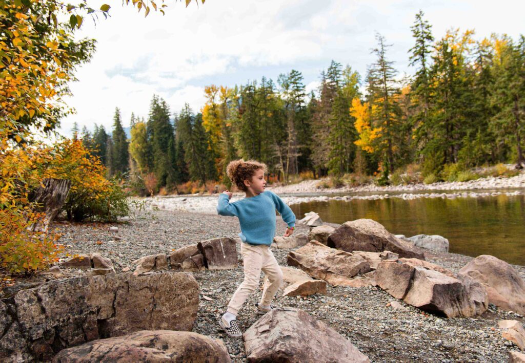 A young child in a blue sweater and light pants stands on a rocky riverbank, mid-motion as they throw a stone towards the water—a perfect moment reminiscent of Spring events at Suncadia, with autumn trees and a calm river in the background.