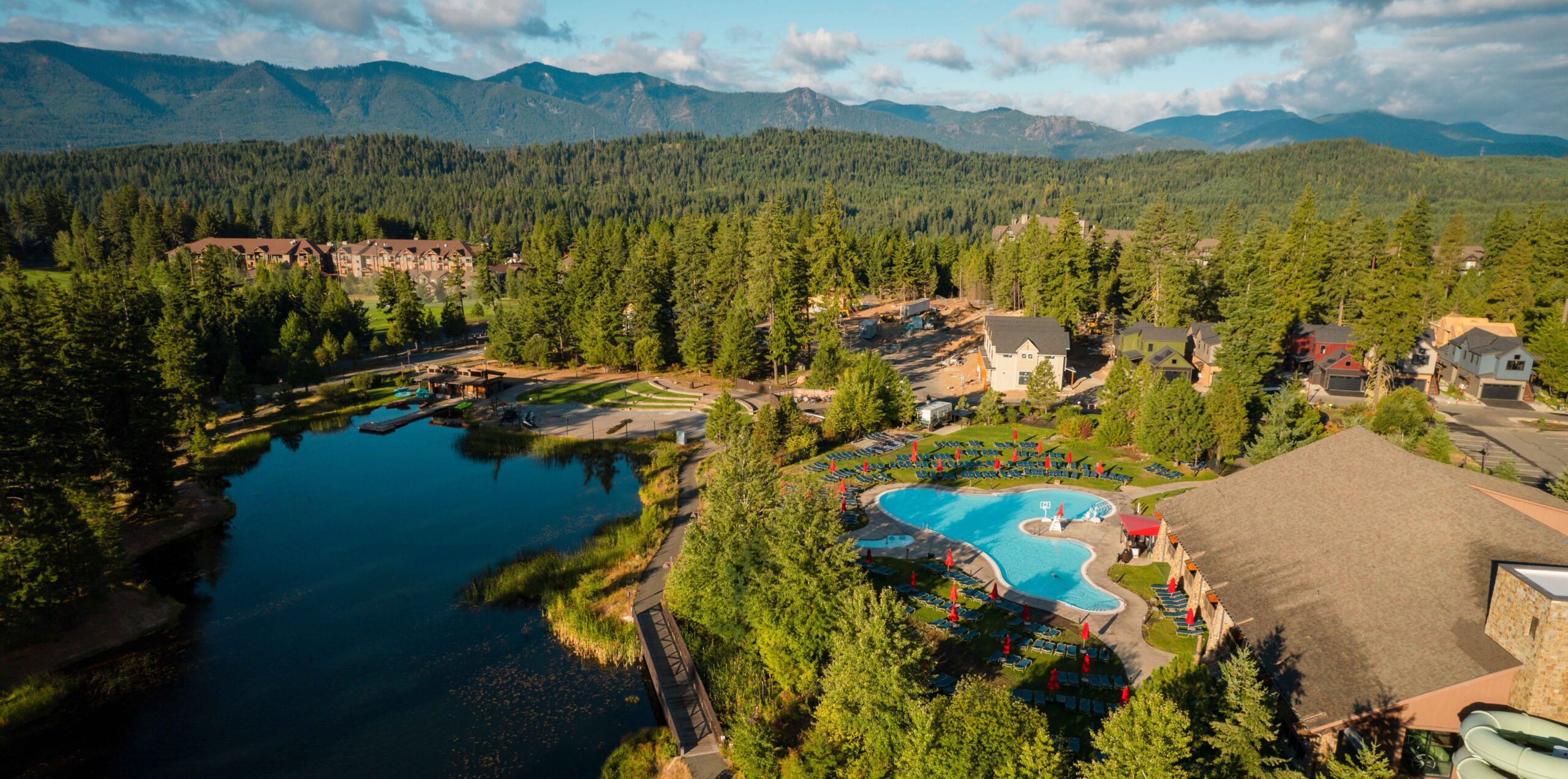 Aerial view of a resort with a large outdoor pool, lounge chairs, and buildings surrounded by dense forest, a small lake, and distant mountains under a partly cloudy sky.