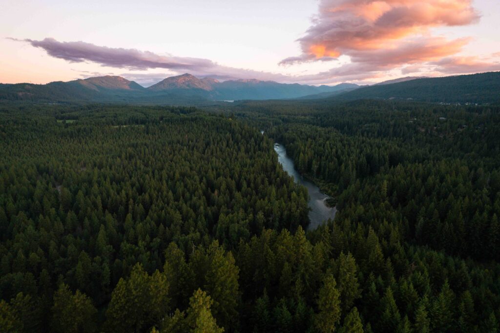 Aerial view of a dense evergreen forest with a river winding through it, set against distant mountains under a sunset sky—an idyllic backdrop for Spring events at Suncadia.