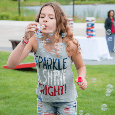 A young girl with long hair blows bubbles outside on a grassy lawn. She wears a tank top that says “SPARKLE & SHINE BRIGHT!” and denim shorts. There are more bubbles and people blurred in the background.
