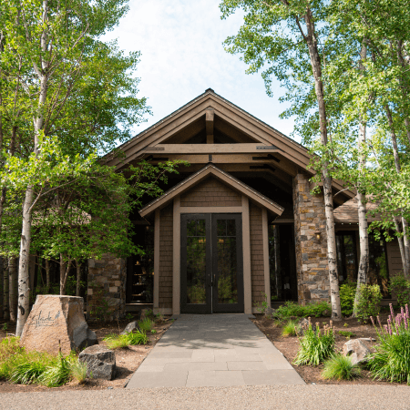 A rustic building with stone and wood exterior surrounded by trees and greenery, featuring a pathway leading to double glass doors at the entrance.