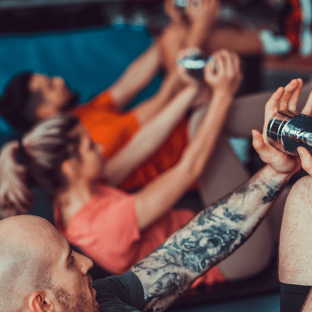 Three people exercising indoors, lying on mats, holding weights with both hands, and raising their arms. The focus is on a tattooed person in the foreground; the others are blurred in the background.