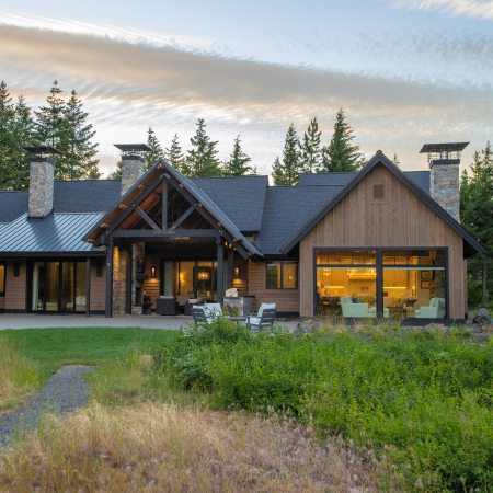 A modern rustic house with large windows, a wooden exterior, and stone chimneys, surrounded by green trees and grass, is pictured at sunset. Outdoor seating is visible on the patio and through the windows.