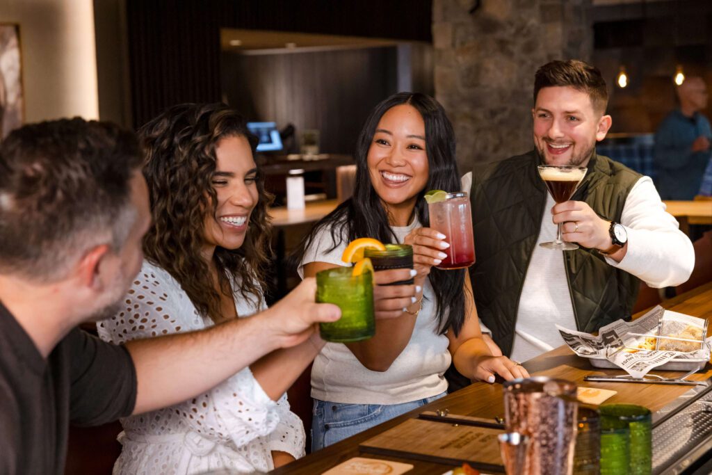 Four friends sit at a restaurant table, smiling and toasting with colorful drinks. They appear happy and engaged, enjoying each other’s company in a cozy, warmly lit setting.