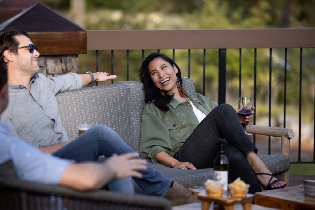 A woman sits on an outdoor couch, smiling and holding a glass of red wine, while talking with two men. There is a table in front of them with a wine bottle and snacks. The setting appears relaxed and social.