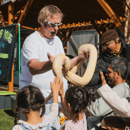 A man wearing sunglasses holds a large yellow and white snake as several children and adults eagerly reach out to touch it during an outdoor event.