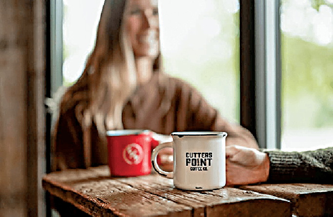 A person sits at a wooden table holding a white mug labeled Cutters Point Coffee Co. Another person, blurred in the background, holds a red mug and smiles near a window.