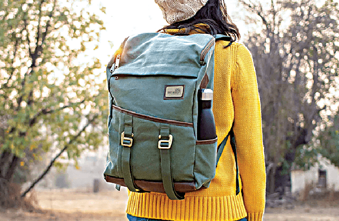 A person wearing a yellow sweater and a knitted hat stands outdoors with a green backpack and a black water bottle attached, surrounded by trees in soft daylight.