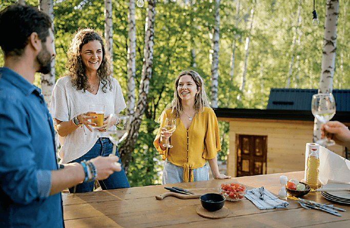 Three people stand outdoors by a wooden table, laughing and holding drinks, with food and drinks on the table. Sunlight shines through trees in the background, creating a relaxed, cheerful atmosphere.