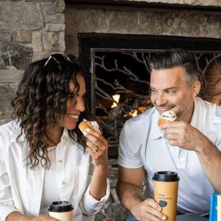 A woman and man sit by a fireplace, smiling at each other while eating pastries. Each holds a coffee cup, and the setting looks cozy and relaxed.