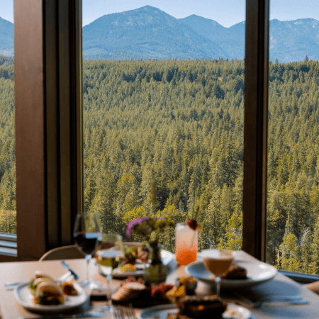 A dining table with assorted food and drinks is set beside large windows, offering a scenic view of a dense green forest and distant mountains under a clear sky.