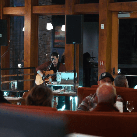 A musician plays an acoustic guitar and sings into a microphone on a small indoor stage, with people seated at tables in the foreground, enjoying the live performance. Warm lighting and wooden decor create a cozy atmosphere.