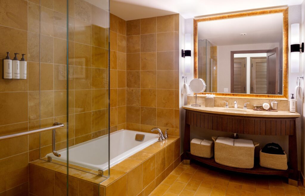 Modern bathroom with tan tiles, a glass-enclosed bathtub, a large mirror above a double sink vanity with two baskets beneath, and wall-mounted lights on either side of the mirror.