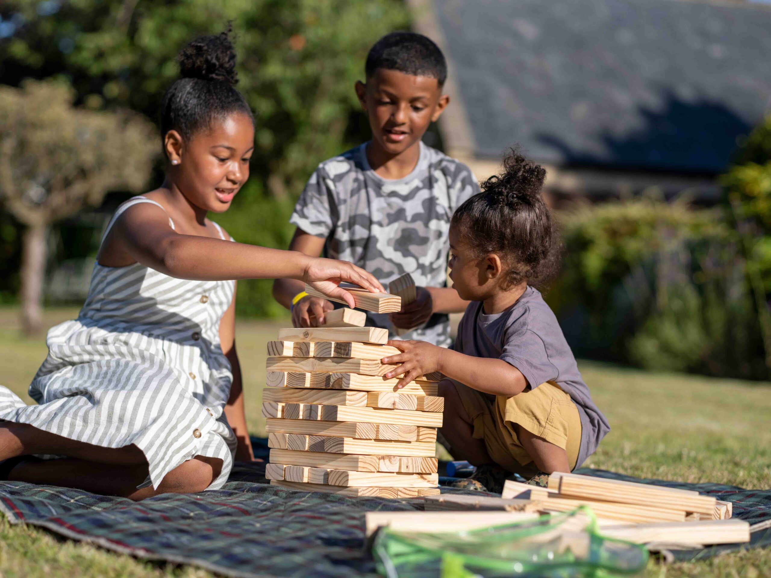 Three children sit on a picnic blanket outdoors, playing a giant wooden block stacking game together on a sunny day. Trees and a building are visible in the background.