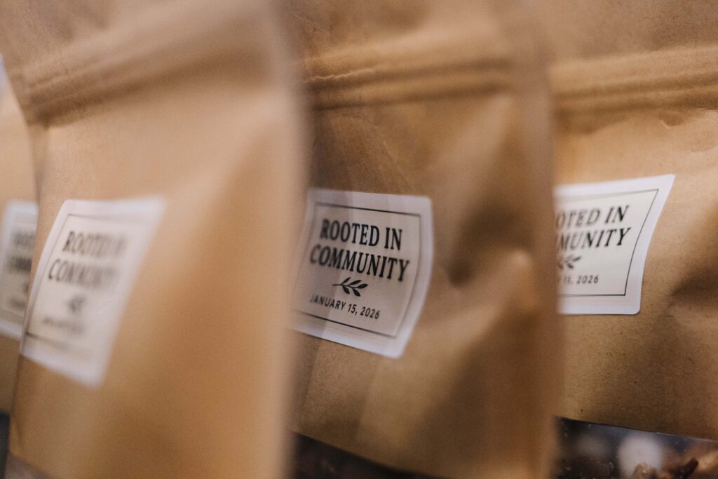 Close-up of brown paper bags with white labels that read Rooted in Community and January 15, 2026. The bags are aligned in a row, partially out of focus.
