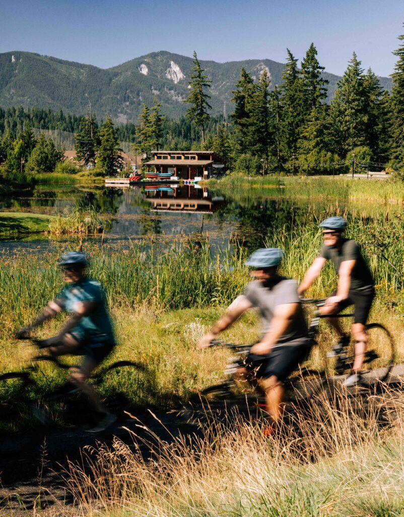 Three cyclists ride along a grassy path in the foreground, appearing blurred from motion. In the background, a wooden house sits by a calm lake, surrounded by trees and mountains under a clear blue sky.