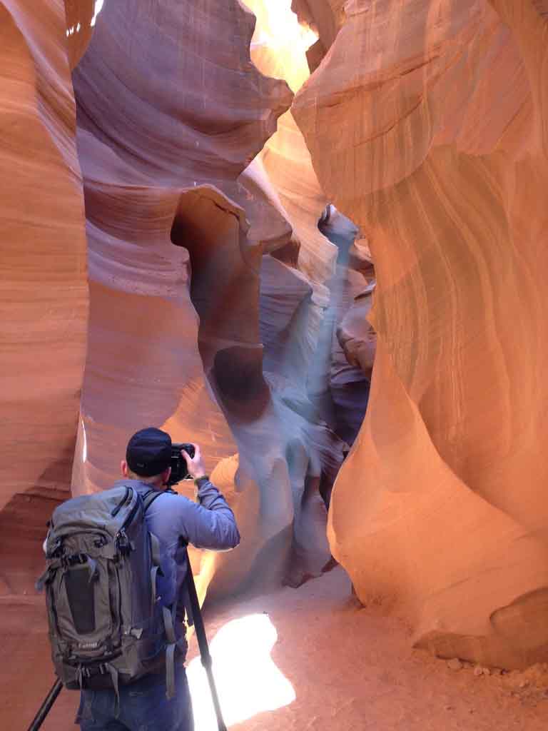 A person with a backpack and camera tripod photographs the narrow, winding sandstone walls of a sunlit slot canyon.