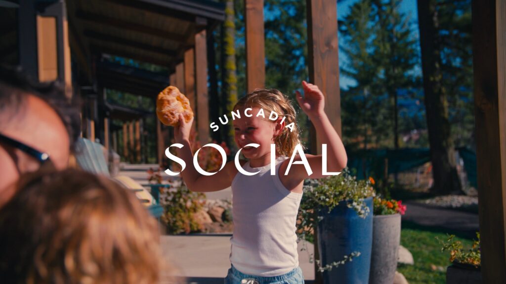 A young child holding a pastry and raising their arms smiles outdoors near a wooden patio, with trees and potted plants in the background. The text Suncadia Social is overlaid on the image.