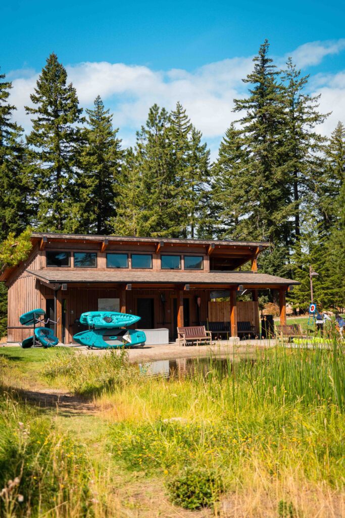A wooden cabin with a covered porch is surrounded by tall evergreen trees and green grass. Several blue kayaks are stacked outside, and two wooden chairs sit on the porch under a bright blue sky.