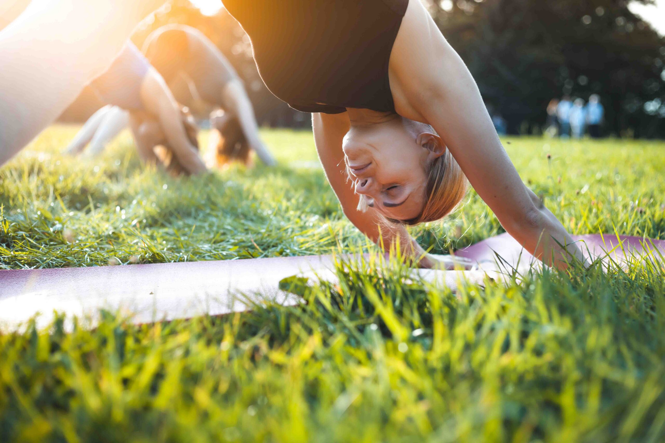 A woman practices yoga outdoors on a grassy field, performing a downward dog pose on a mat, with other people in the background doing the same. Sunlight filters through the trees.