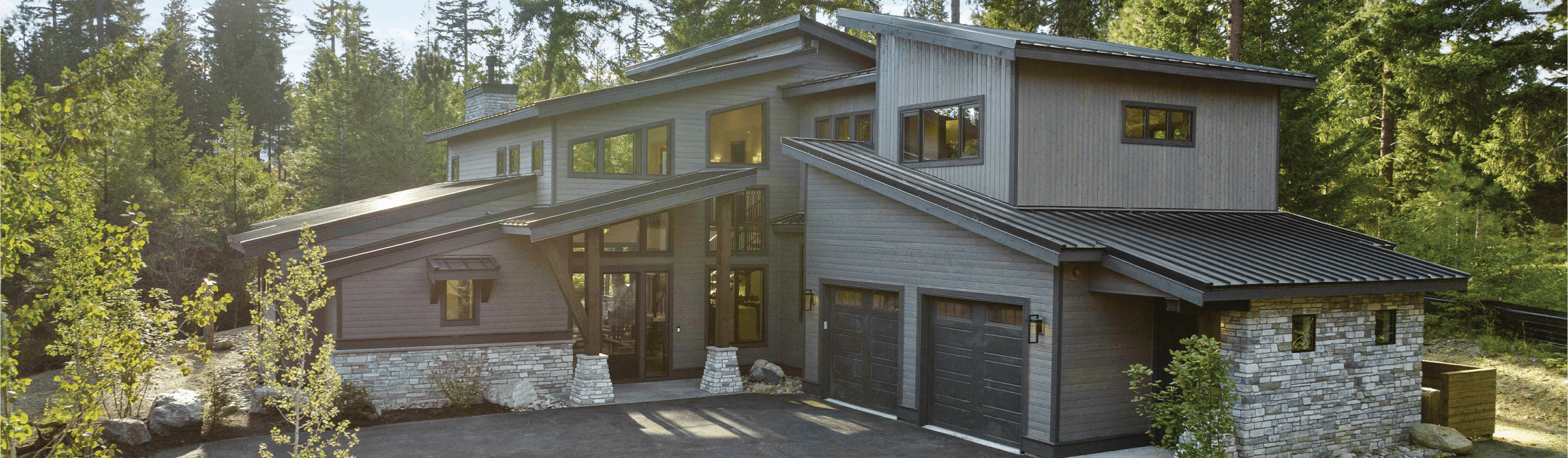 Modern two-story house with dark wood siding, large windows, and a stone accent exterior, surrounded by trees. The driveway leads to a two-car garage with dark doors.