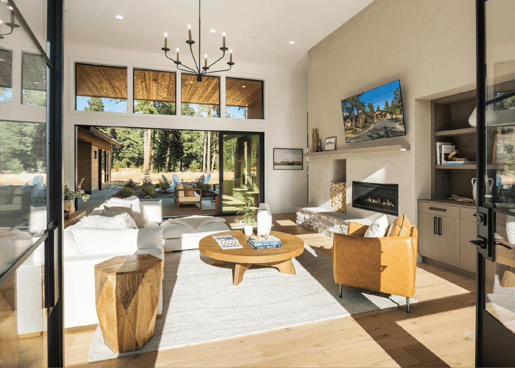 Bright, modern living room with large windows, white sofas, tan leather chair, round wooden coffee table, fireplace, built-in shelves, and a view of a patio and trees outside. Neutral decor and natural light fill the space.