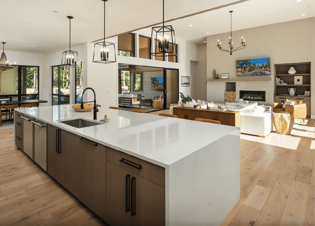 Modern open-concept kitchen and living area with a large white island, wooden cabinets, hanging lights, cozy seating, fireplace, and large windows overlooking trees. Light wood floors throughout.