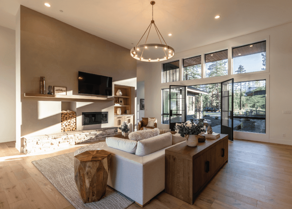 Modern living room with large windows, neutral decor, a white sofa, wooden coffee table, fireplace with stacked firewood, wall-mounted TV, and a circular chandelier. Sunlight pours in from glass doors showing green trees outside.