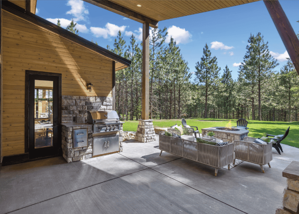 Covered patio with outdoor kitchen, stainless steel grill, and modern seating area, overlooking a lawn and pine forest under a blue sky with scattered clouds.