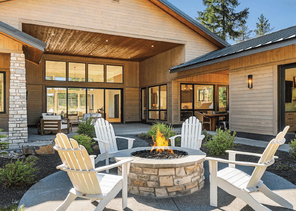 An outdoor patio with four white Adirondack chairs arranged around a stone fire pit, in front of a modern house with large windows and wooden siding, surrounded by greenery.