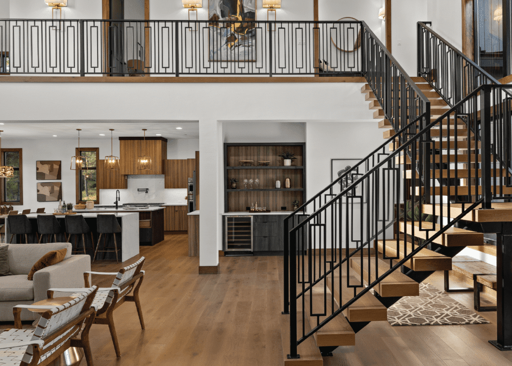 Modern open-concept living space with wooden floors, a staircase with black railings, a kitchen with wood cabinets and white countertops, a wine fridge, and stylish furniture, all brightly lit by natural and pendant lighting.