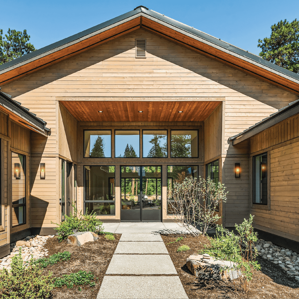 A modern wooden building with large glass doors and windows, a peaked roof, and exterior lights. A stone pathway leads to the entrance, bordered by landscaped plants and mulch under a clear blue sky.