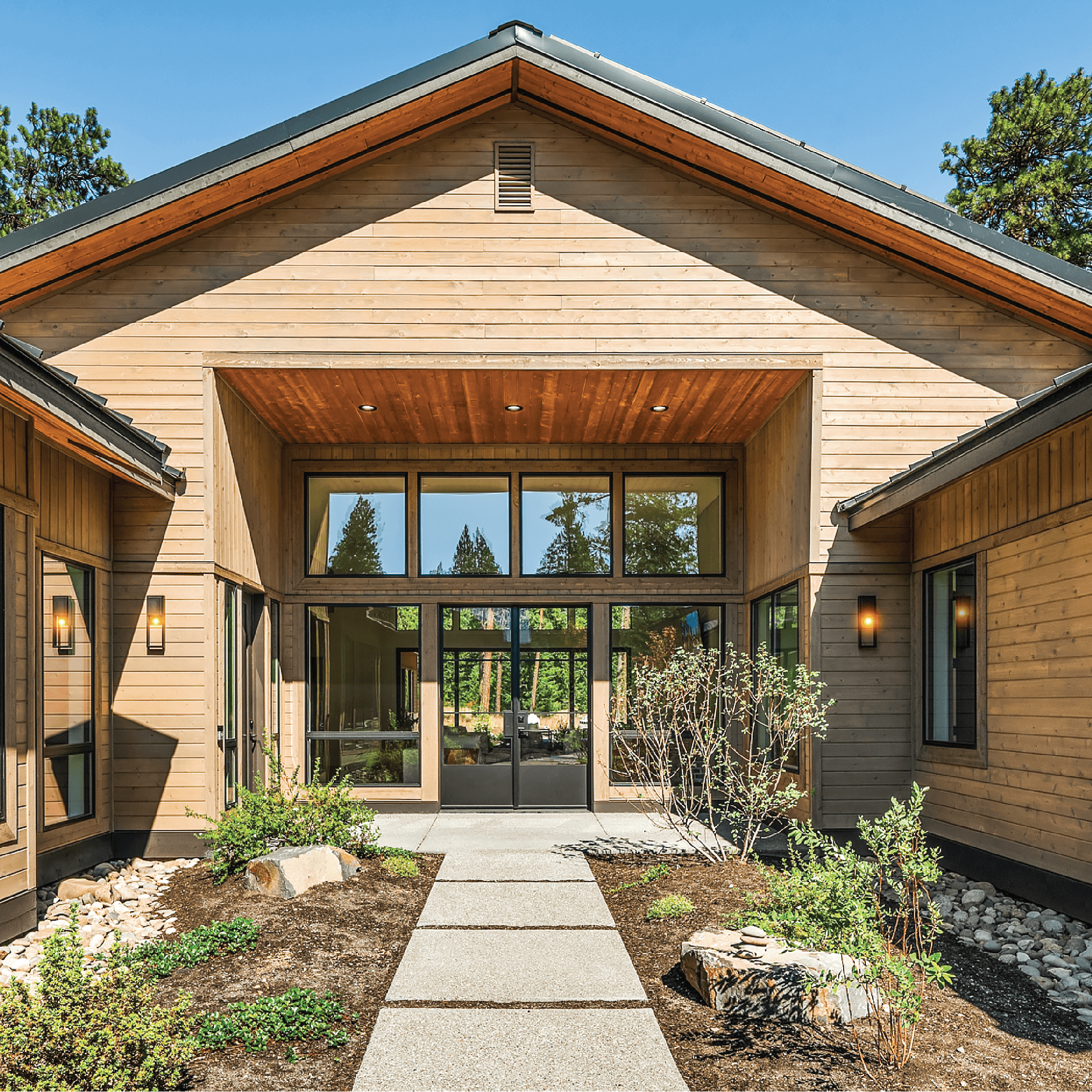 A modern wooden building with large glass doors and windows, a peaked roof, and exterior lights. A stone pathway leads to the entrance, bordered by landscaped plants and mulch under a clear blue sky.