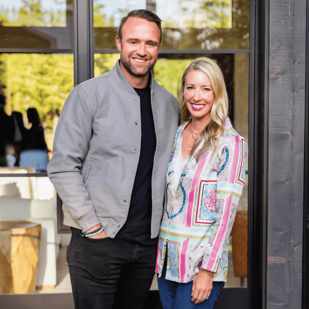 A man in a gray jacket and black shirt stands next to a woman in a colorful patterned blouse. They are both smiling and posing outside in front of a glass door with greenery reflected in the background.