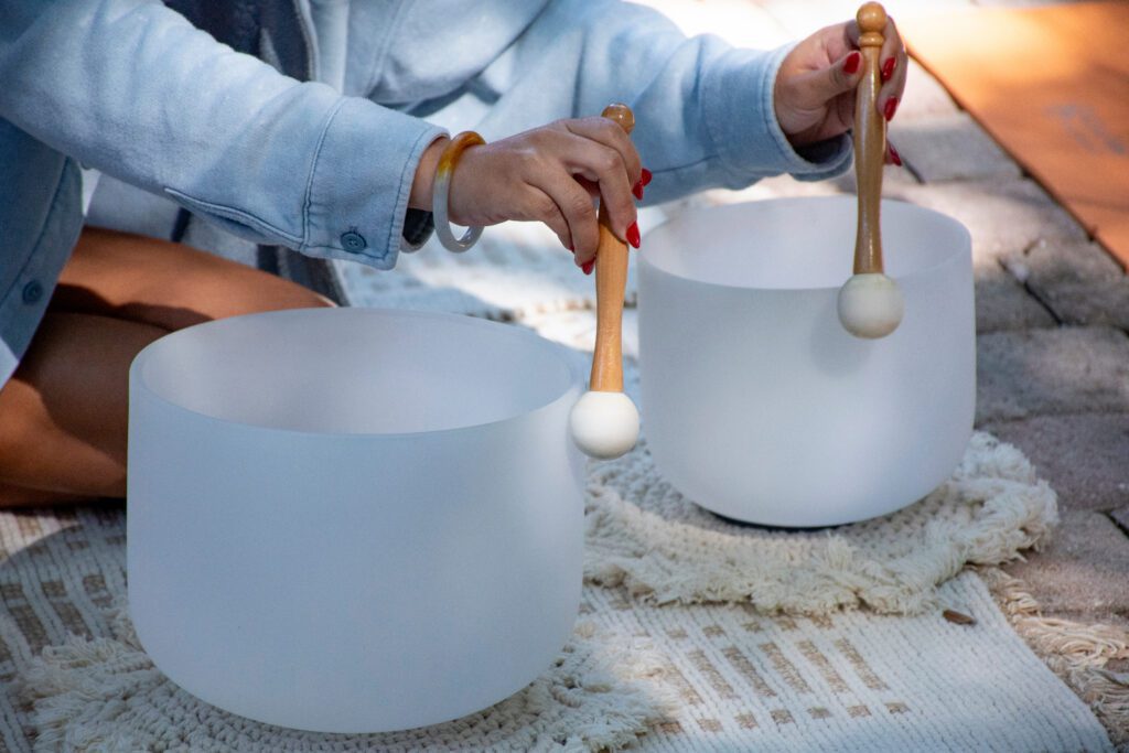 A person uses two mallets to play a pair of white crystal singing bowls placed on a patterned rug outdoors. Only their hands and part of their body are visible.