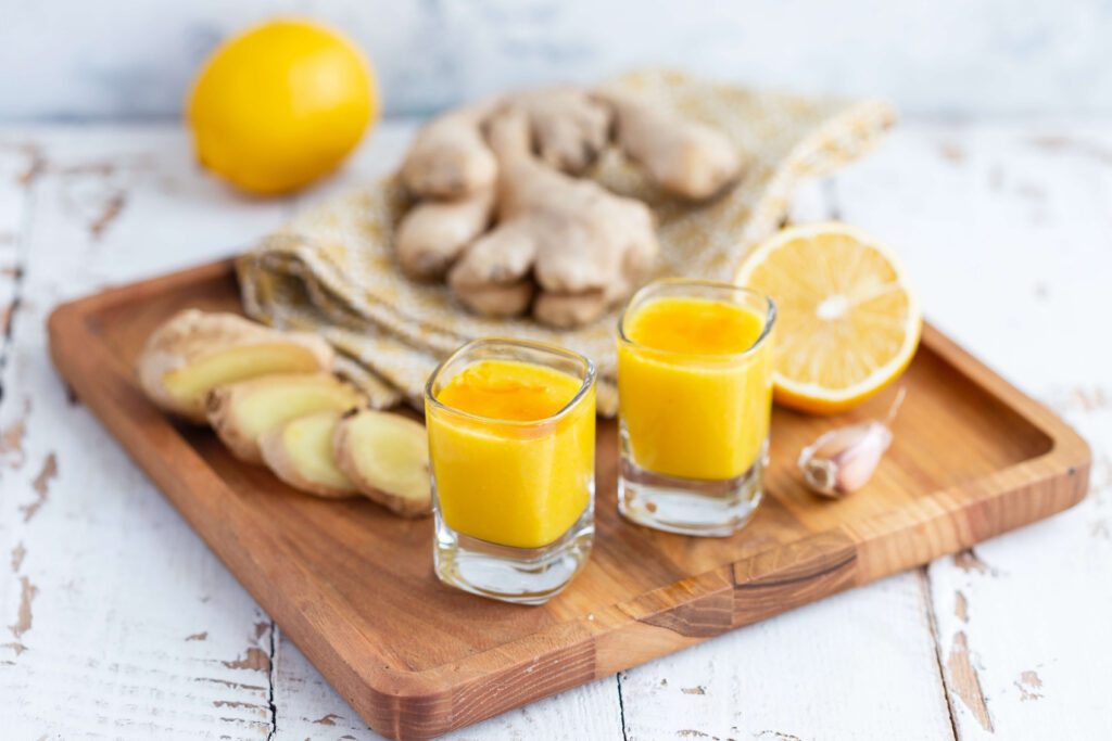 Two small glasses of yellow juice sit on a wooden tray with ginger slices, a halved lemon, a garlic clove, a whole lemon, and ginger root in the background on a white rustic surface.