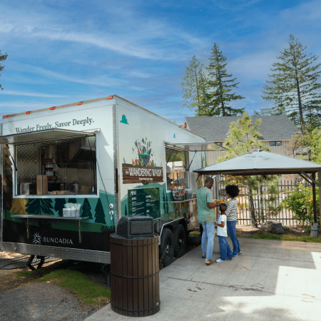 A family stands by a green food truck labeled “Wandering Wafer,” parked near trees and a small covered seating area on a sunny day. A trash bin is beside the truck, and homes are visible in the background.