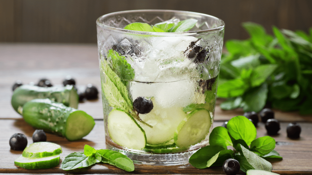 A glass of iced water with cucumber slices, fresh mint leaves, and blueberries, sits on a wooden table with more cucumbers, mint, and blueberries scattered around.