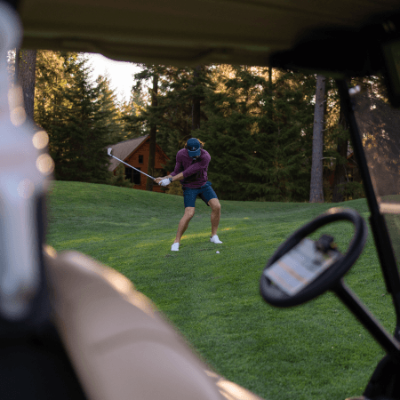 A person wearing a cap, purple shirt, and shorts swings a golf club on a grassy course, viewed through a golf cart with trees and cabins in the background.