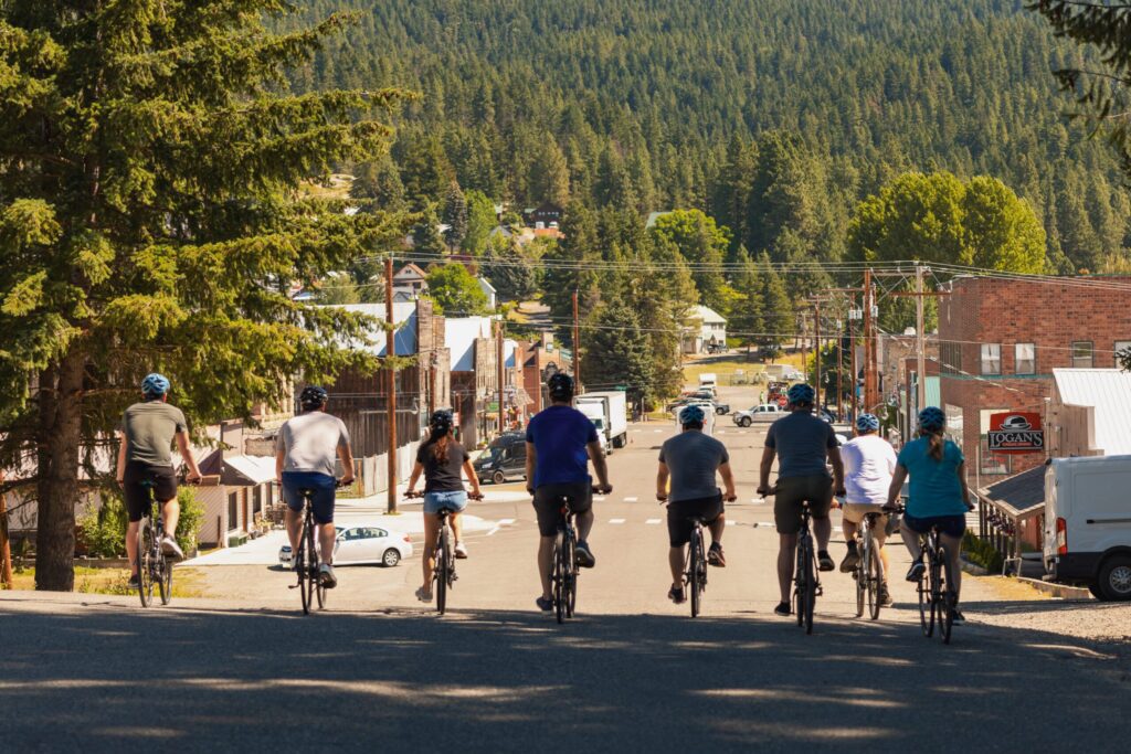 A group of eight people wearing helmets ride bicycles down a street toward a small town, surrounded by trees and forested hills on a sunny day.