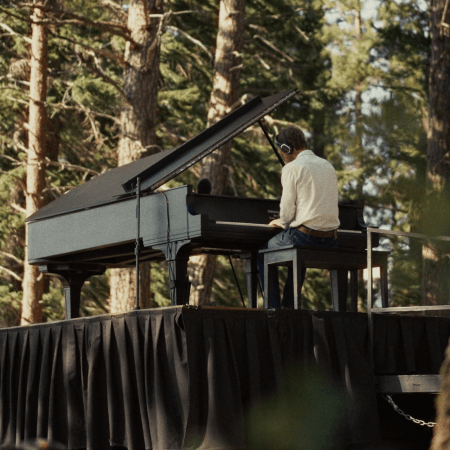 A person wearing headphones plays a grand piano on an outdoor stage surrounded by tall trees and sunlight, with a black tablecloth draped over the stage.