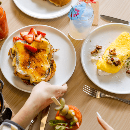 Overhead view of a brunch table with stuffed French toast topped with strawberries, an omelette with potatoes, beverages including a cocktail and a drink with an umbrella, and a person reaching for food.