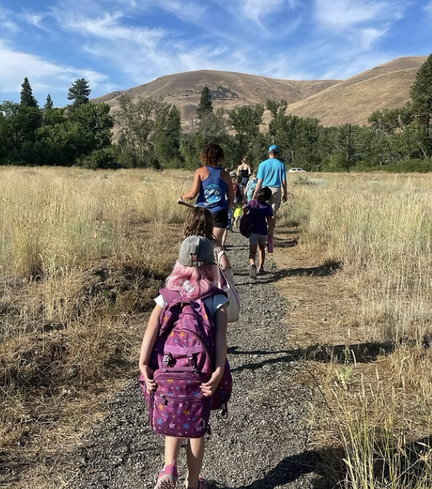 A group of people, including children with backpacks, walk single file along a dirt trail through dry grass, heading toward hills under a blue sky with wispy clouds.