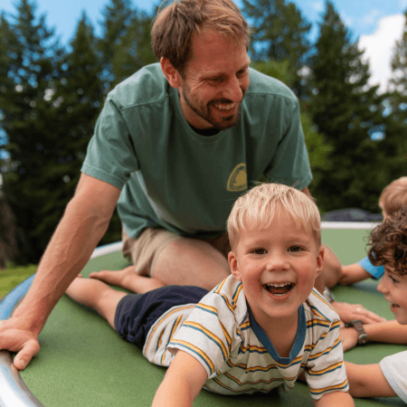 A smiling man and young children play together outdoors on a trampoline, surrounded by trees and blue sky. The boy in the front is laughing and lying on his stomach, while the others enjoy the activity.