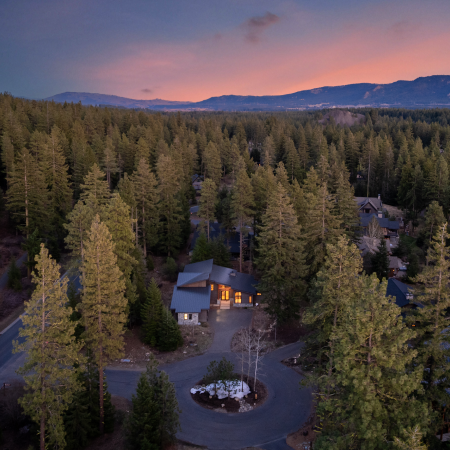 A modern house surrounded by tall pine trees is illuminated at dusk, with winding roads and mountains visible in the background under a colorful sunset sky.
